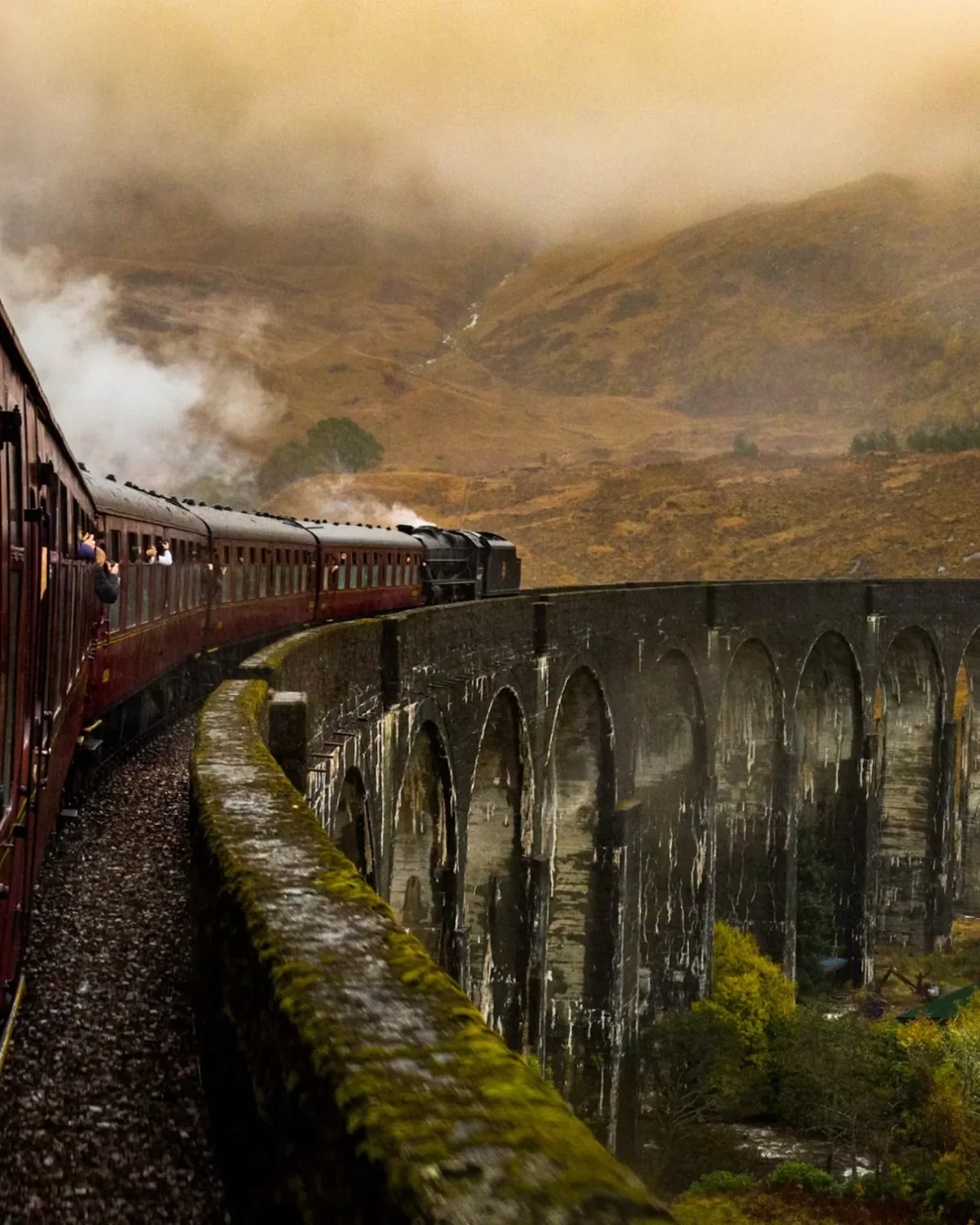 A red steam train crossing a stone viaduct in misty highlands